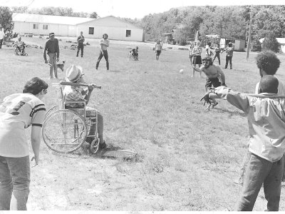 A young man pitches a ball to a boy sitting in a wheelchair and holding a bat. Other children and young adults look ready to react.