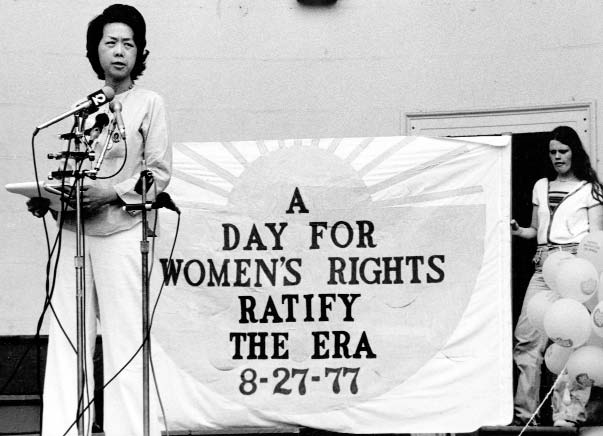 A Chinese American woman speaks into multiple microphones at a rally. A sign behind her reads "A Day for Women's Rights. Ratify the ERA (Equal Rights Amendment) 8-27-77"