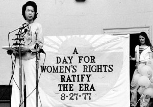 A Chinese American woman speaks into multiple microphones at a rally. A sign behind her reads "A Day for Women's Rights. Ratify the ERA (Equal Rights Amendment) 8-27-77"