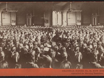 A stereoscophic slide with photos taken from two slightly different angles shows around 100 schoolgirls seated in a large auditorium with several women teachers in the background.