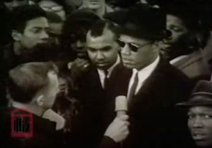 Malcolm X is interviewed while standing in front of a crowd of people.