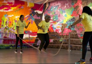 Reverand Malika Leigh Whitney, an older black woman, jumps rope with two women swinging two ropes.