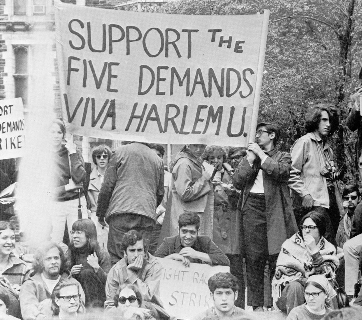 College students hold signs at an outdoor rally. A large banner reads, "Support the Five Demands - Viva Harlem U."