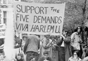 College students hold signs at an outdoor rally. A large banner reads, "Support the Five Demands - Viva Harlem U."